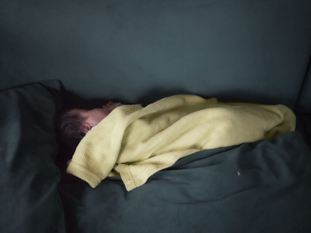Photographie de Zélie Hallosserie / Light Motiv, issue de la série intitulée The Game, représentant un bébé qui dort enveloppé d'une couverture jaune posé sur ce qui ressemble à un lit couvert de draps foncés, dans le cadre d'un reportage documentaire porté sur le voyage des migrants dans le nord de la France, sur la côte.