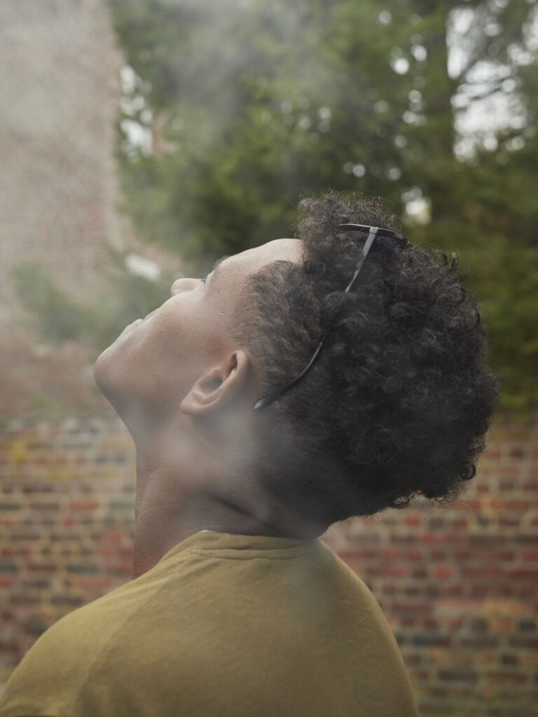 Photographie de Zélie Hallosserie / Light Motiv, issue de la série intitulée The Game, représentant un plan rapproché d'un homme regardant vers le coin supérieur gauche du cadre de l'image d'où se répand une fumée blanche, dans le cadre d'un reportage documentaire porté sur le voyage des migrants dans le nord de la France, sur la côte.