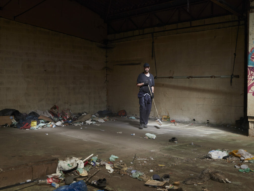Photographie de Zélie Hallosserie / Light Motiv, issue de la série intitulée The Game, représentant un homme debout avec un balais dans un intérieur sombre dont le sol est recouvert de déchets, dans le cadre d'un reportage documentaire porté sur le voyage des migrants dans le nord de la France, sur la côte.