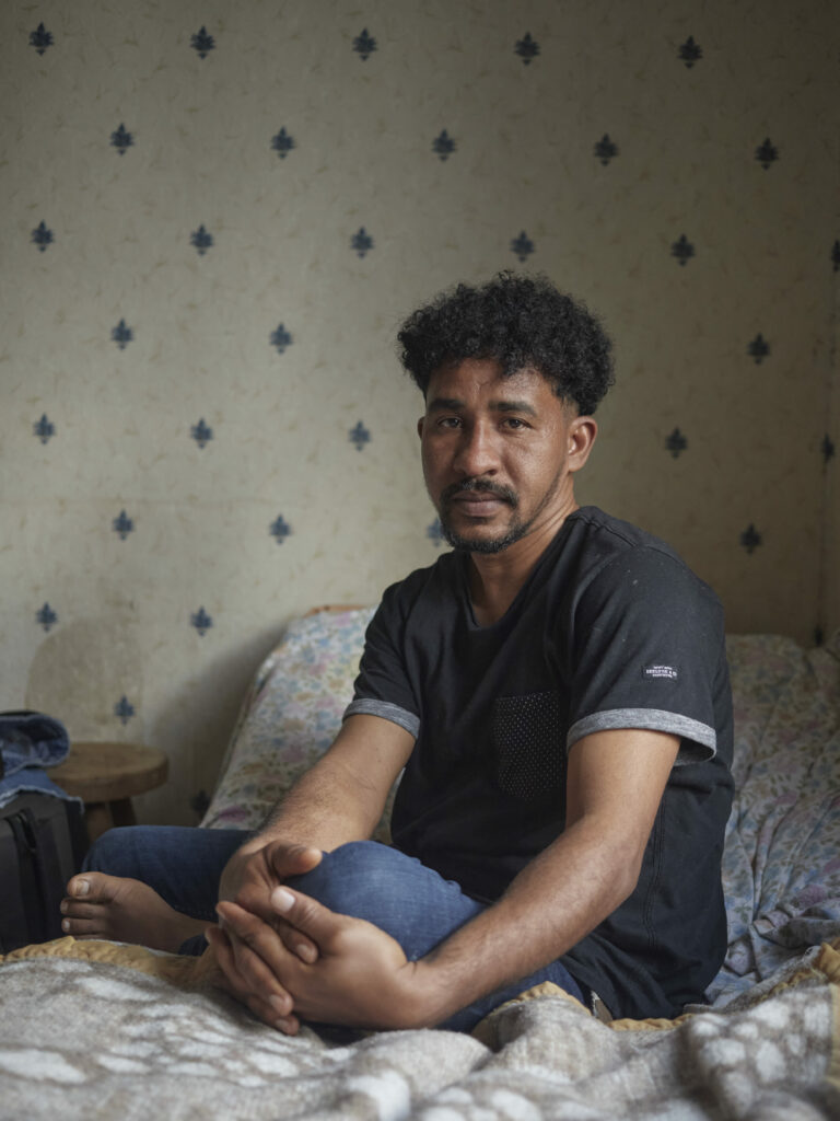 Photographie de Zélie Hallosserie / Light Motiv, issue de la série intitulée The Game, représentant un homme dans un intérieur assis sur un lit dans une posture décontracté qui regarde vers la caméra, dans le cadre d'un reportage documentaire porté sur le voyage des migrants dans le nord de la France, sur la côte.