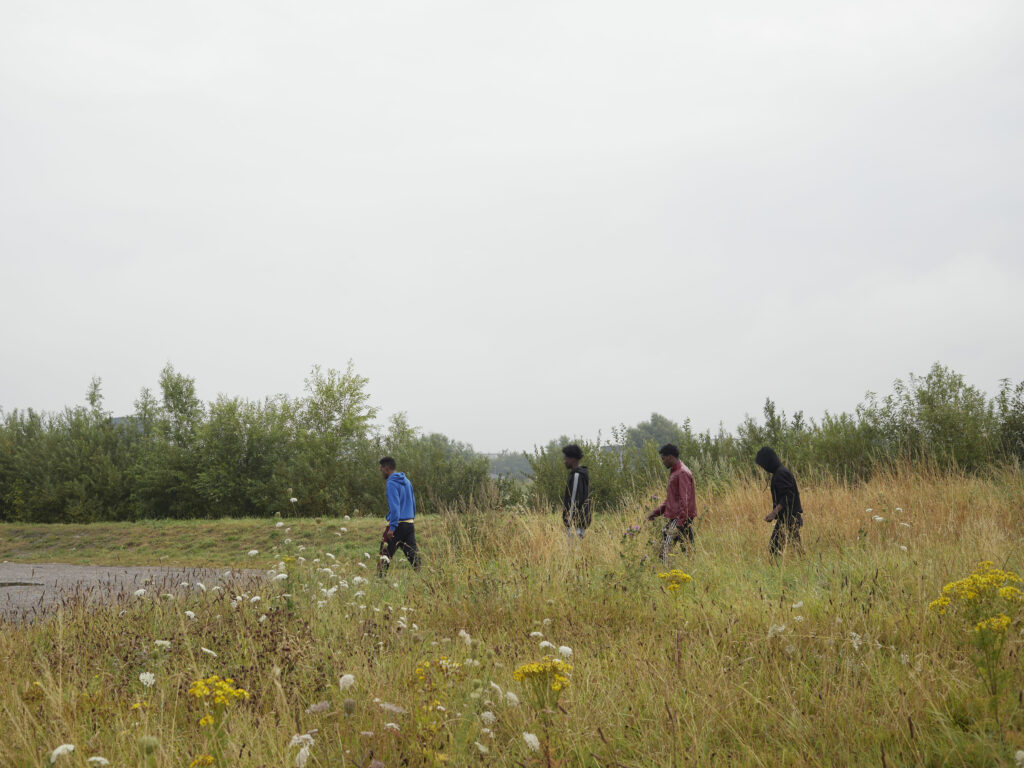 Photographie de Zélie Hallosserie / Light Motiv, issue de la série intitulée The Game, représentant un groupe de quatres hommes vu de loin dans un terrain végétalisé marchant vers la gauche de l'image, dans le cadre d'un reportage documentaire porté sur le voyage des migrants dans le nord de la France, sur la côte.