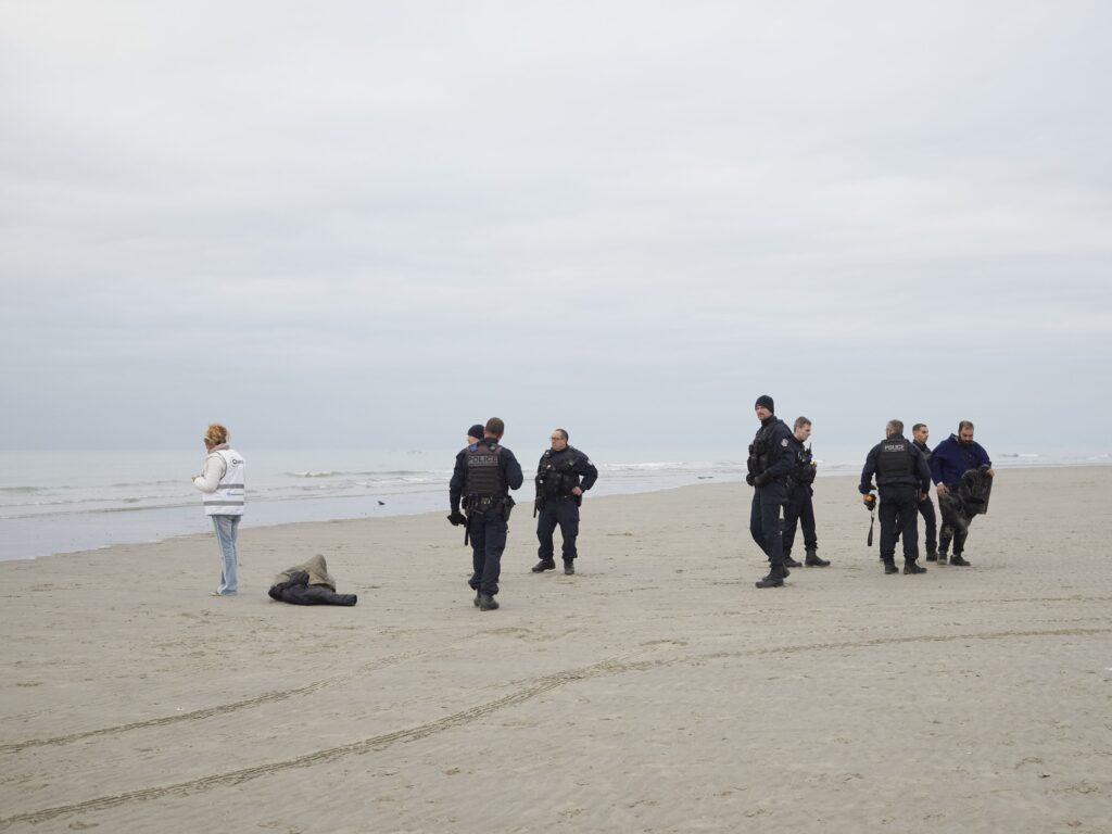 Photographie de Zélie Hallosserie / Light Motiv, issue de la série intitulée The Game, représentant un groupe de policiers sur une plage à proximité de la mer regardant autour d'eux, dans le cadre d'un reportage documentaire porté sur le voyage des migrants dans le nord de la France, sur la côte.