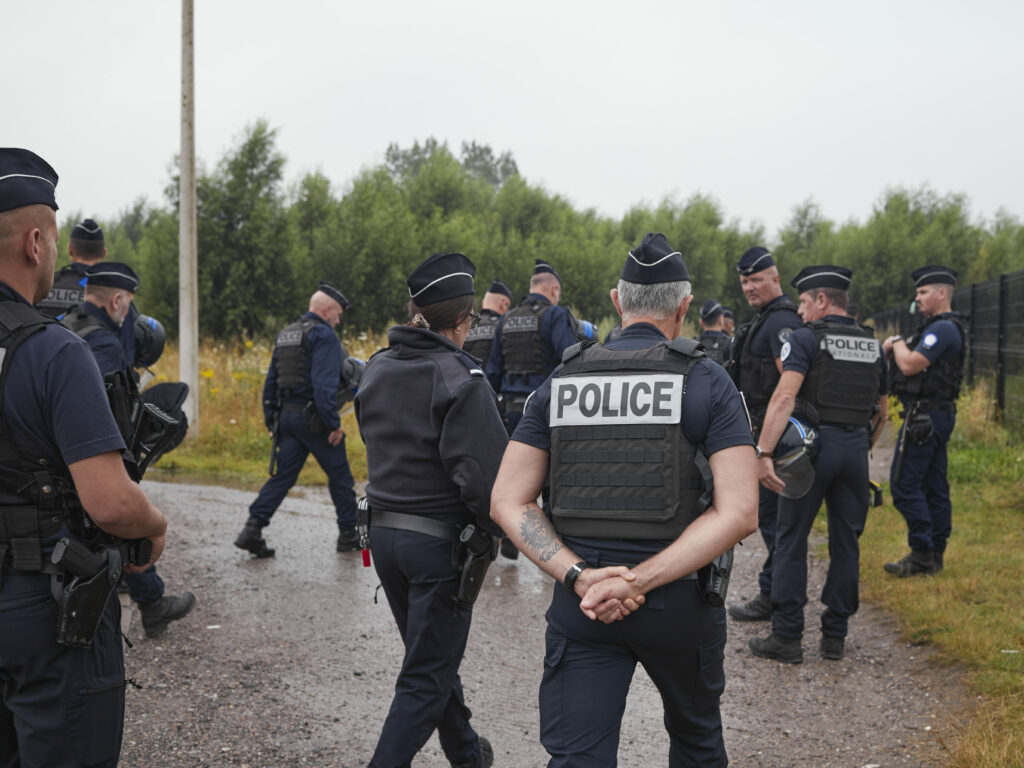 Photographie de Zélie Hallosserie / Light Motiv, issue de la série intitulée The Game, représentant un groupe de policiers qui marche sur un terrain végétalisé, dans le cadre d'un reportage documentaire porté sur le voyage des migrants dans le nord de la France, sur la côte.