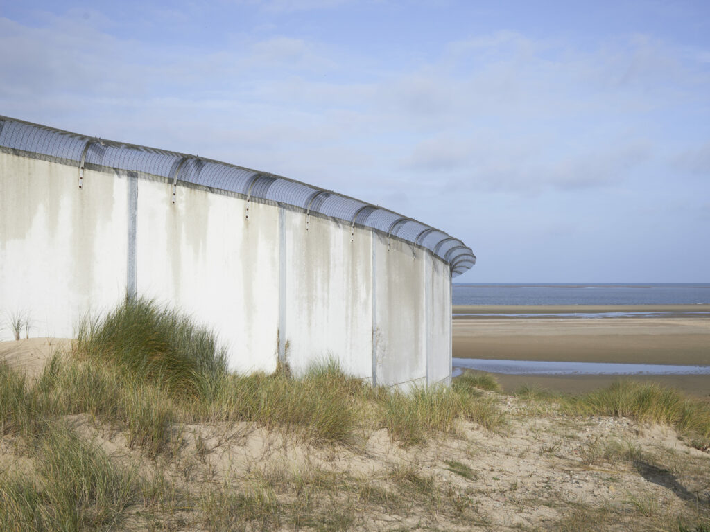 Photographie de Zélie Hallosserie / Light Motiv, issue de la série intitulée The Game, représentant un mur avec un grillage courbé vers l'extérieur à son sommet dans des dunes à proximité d'une plage, dans le cadre d'un reportage documentaire porté sur le voyage des migrants dans le nord de la France, sur la côte.