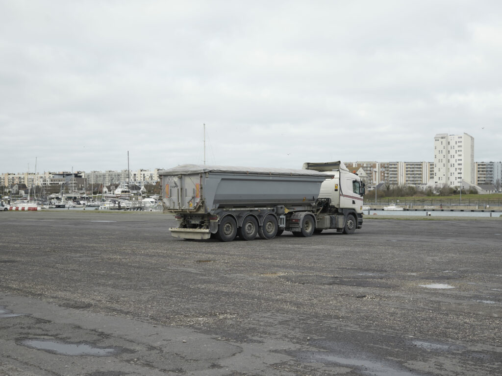 Photographie de Zélie Hallosserie / Light Motiv, issue de la série intitulée The Game, représentant un camion à l'arrêt sur un grand parking aux abords d'un port, dans le cadre d'un reportage documentaire porté sur le voyage des migrants dans le nord de la France, sur la côte.