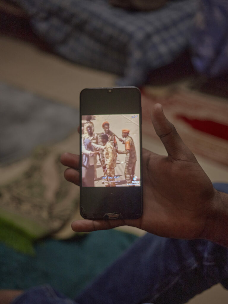 Photographie de Zélie Hallosserie / Light Motiv, issue de la série intitulée The Game, représentant un gros plan sur un téléphone tenu dans une main qui présente une vidéo TikTok sur des militaires, dans le cadre d'un reportage documentaire porté sur le voyage des migrants dans le nord de la France, sur la côte.