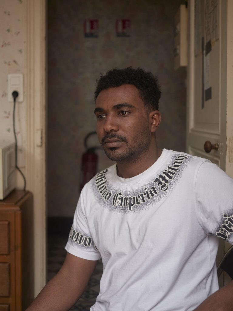 Photographie de Zélie Hallosserie / Light Motiv, issue de la série intitulée The Game, représentant un homme assis dans un intérieur au t-shirt blanc qui regarde à côté de la caméra d'un air perdu, dans le cadre d'un reportage documentaire porté sur le voyage des migrants dans le nord de la France, sur la côte.