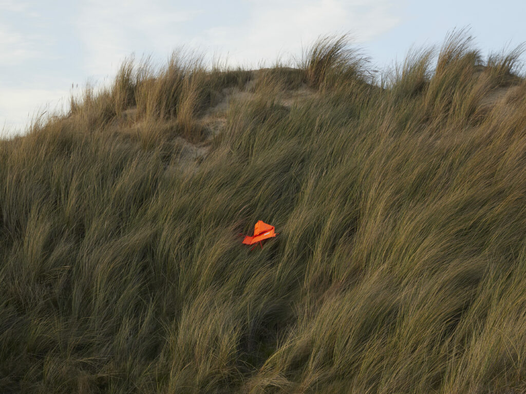 Photographie de Zélie Hallosserie / Light Motiv, issue de la série intitulée The Game, représentant une vue dans des dunes avec un gilet de sauvetage orange posé dans les hautes herbes, dans le cadre d'un reportage documentaire porté sur le voyage des migrants dans le nord de la France, sur la côte.