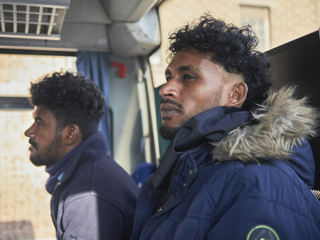 Photographie de Zélie Hallosserie / Light Motiv, issue de la série intitulée The Game, représentant un plan poitrine de deux hommes assis dans un bus qui regardent vers la gauche (à l'avant du bus), dans le cadre d'un reportage documentaire porté sur le voyage des migrants dans le nord de la France, sur la côte.