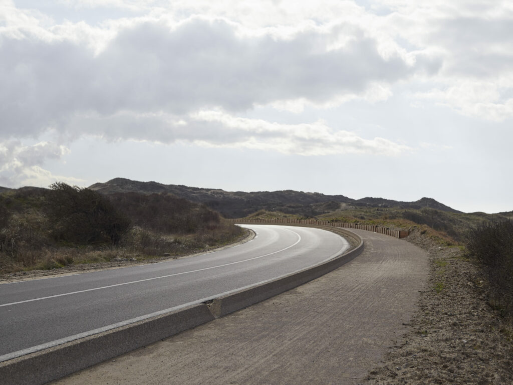 Photographie de Zélie Hallosserie / Light Motiv, issue de la série intitulée The Game, représentant une route aux abords des dunes qui se poursuit dans un virage, dans le cadre d'un reportage documentaire porté sur le voyage des migrants dans le nord de la France, sur la côte.