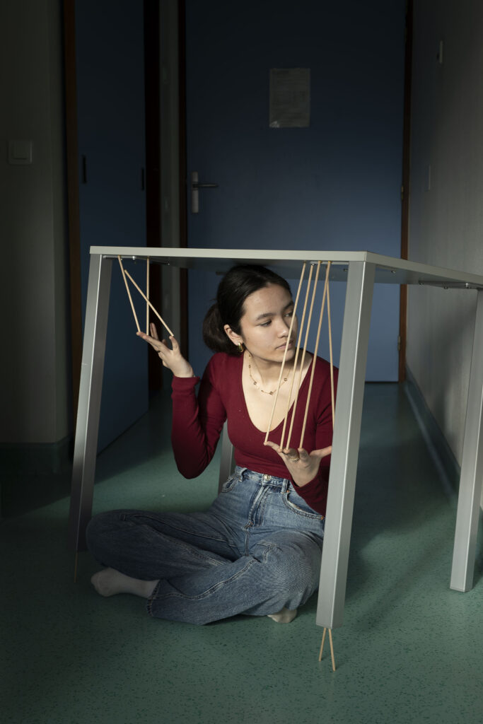 Photographie de Lionel Pralus issue de sa série intitulée "dés-équilibres" représentant le portrait d'une jeune personne assise sur le sol, supportant une table métallique au-dessus d'elle avec des bouts de bois tenus entre ses doigts et le plateau de la table. Lors d'une résidence en 2024 au sein du Service Adolescents  de l'Hôpital St Vincent de Paul de Lille.