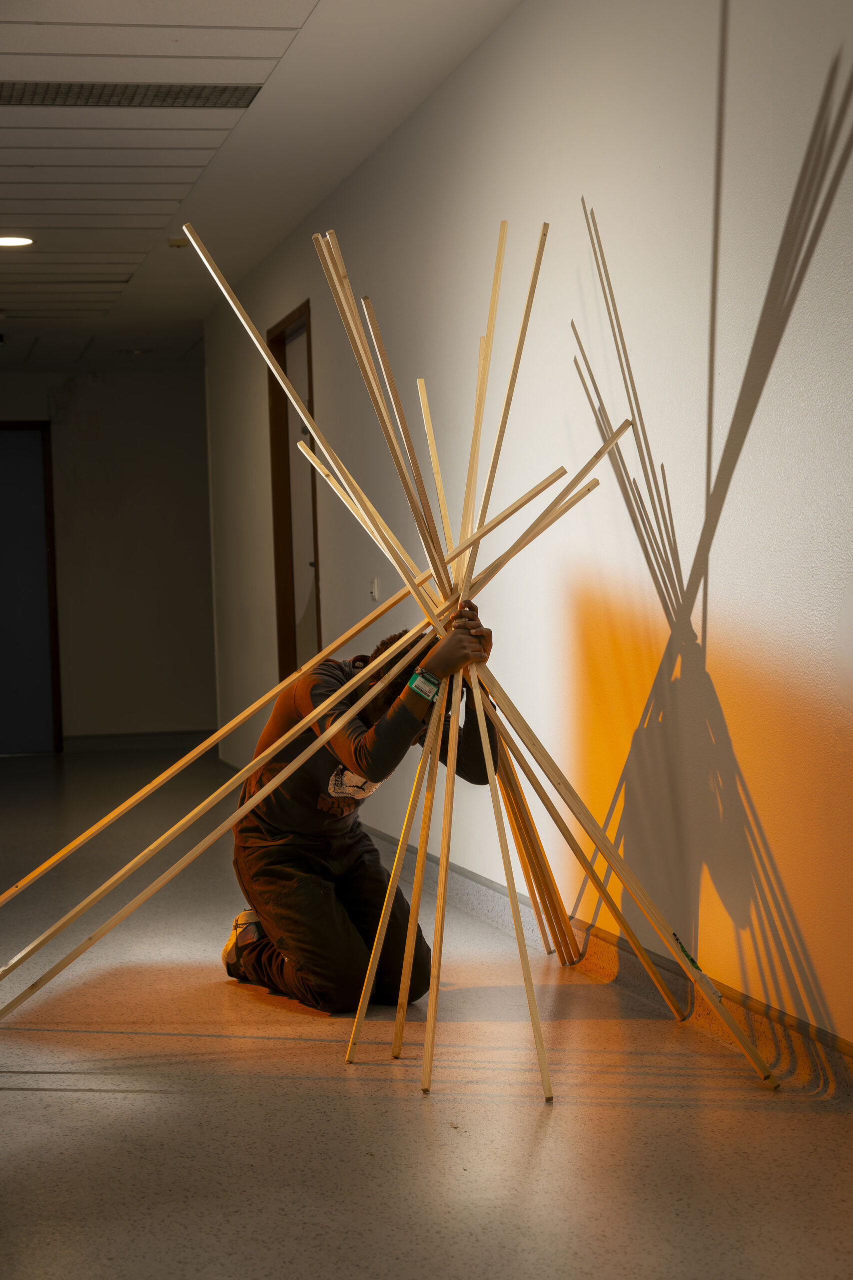 Photographie de Lionel Pralus issue de sa série intitulée "dés-équilibres" représentant le portrait d'une personne accroupie sur le sol dans un couloir faiblement éclairé, tenant dans ses mains une grande structure en bois en forme de tipi. Lors d'une résidence en 2024 au sein du Service Adolescents de l'Hôpital St Vincent de Paul de Lille.