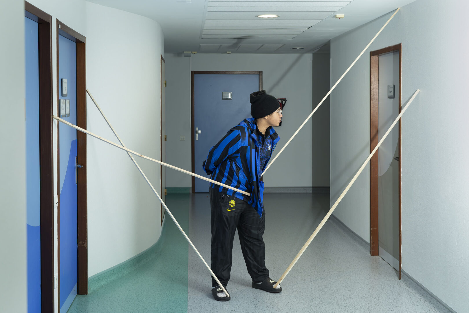 Photographie de Lionel Pralus issue de sa série intitulée "dés-équilibres" représentant le portrait en pied d'une personne debout dans un couloir, appuyée sur 4 longues tiges de bois supportées sur les murs annexes. Lors d'une résidence en 2024 au sein du Service Adolescents de l'Hôpital St Vincent de Paul de Lille.