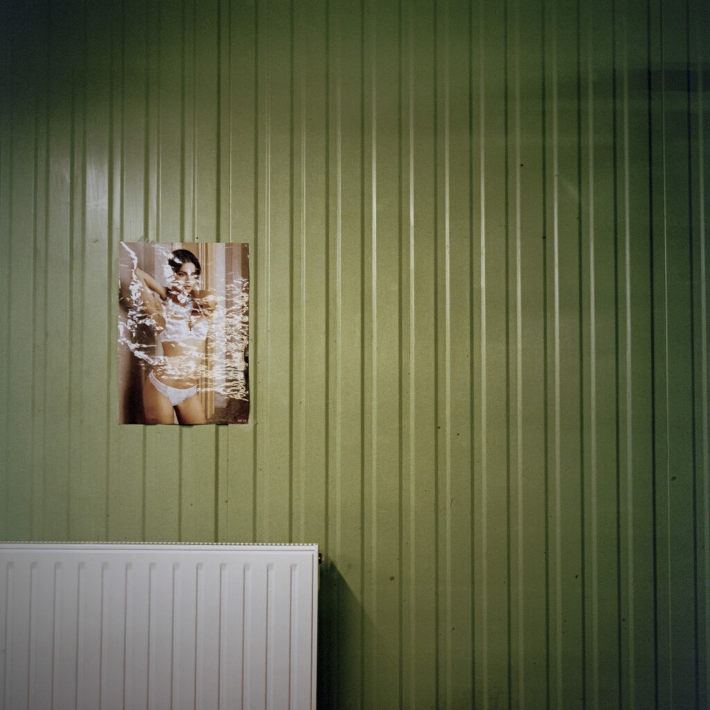 Photographie de Zélie Hallosserie / Light Motiv, issue de la série intitulée Dentelle Calais Caudry, représentant une image, représentant une femme en lingerie de dentelles blanches, accrochée sur un mur vert olive avec un radiateur blanc dans le coin inférieur gauche de l'image, dans le cadre d'un reportage qui témoigne du travail et de la conception de la dentelle à Calais.