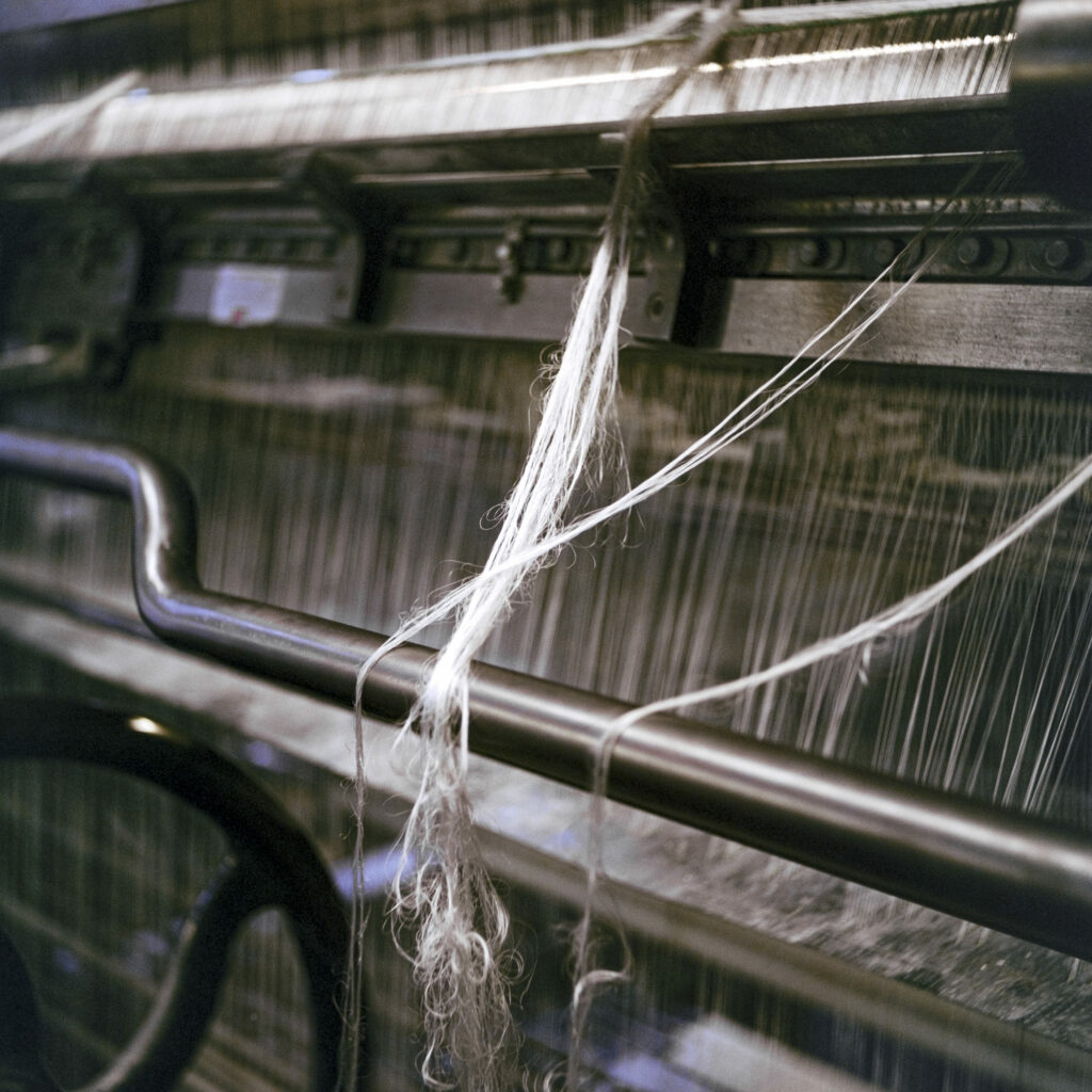 Photographie de Zélie Hallosserie / Light Motiv, issue de la série intitulée Dentelle Calais Caudry, représentant un gros plan d'un métier à tisser de la dentelle dont des fils blancs pendent en travers de celui-ci, dans le cadre d'un reportage qui témoigne du travail et de la conception de la dentelle à Calais.