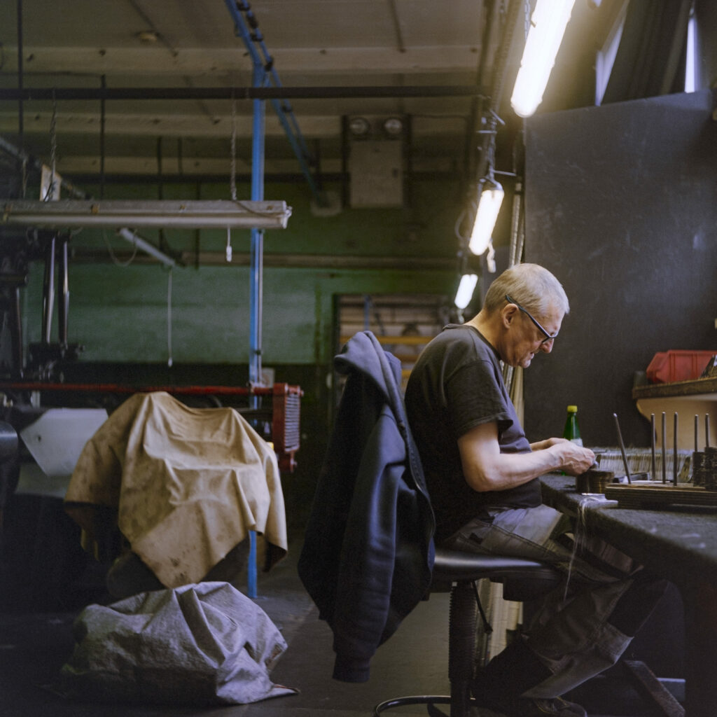 Photographie de Zélie Hallosserie / Light Motiv, issue de la série intitulée Dentelle Calais Caudry, représentant un homme assis sur une chaise dans un atelier occupé avec ses mains sur un plan de travail sur la droite de l'image, dans le cadre d'un reportage qui témoigne du travail et de la conception de la dentelle à Calais.
