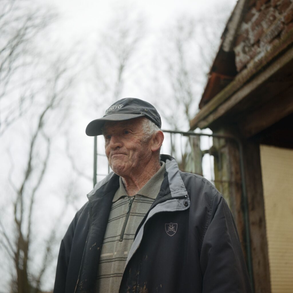 Photographie de Zélie Hallosserie / Light Motiv, issue de la série intitulé Jean Marie, représentant un plan poitrine de Jean Marie debout à l'extérieur regardant au loin vers la gauche de l'image , dans l'intention de capturer les moments de vie de Jean Marie.