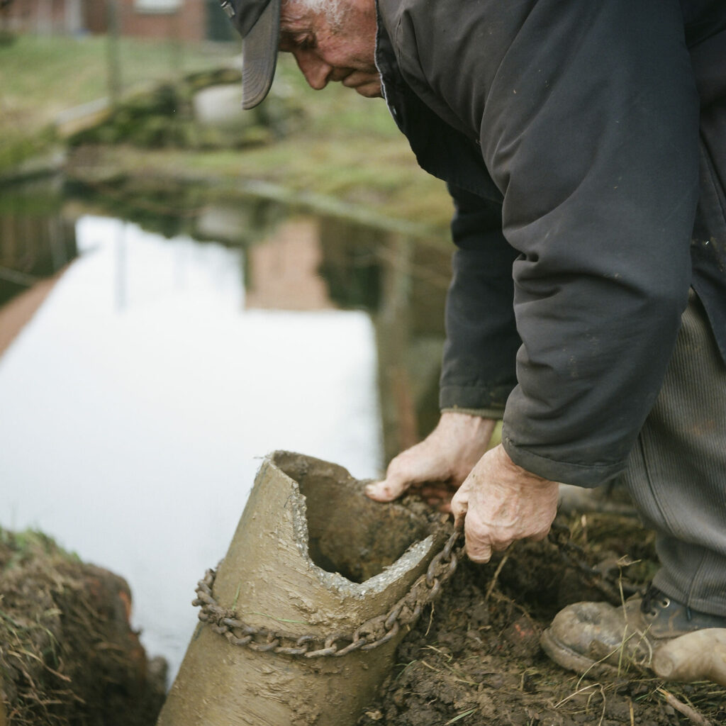 Photographie de Zélie Hallosserie / Light Motiv, issue de la série intitulé Jean Marie, représentant Jean Marie soulevant avec ses mains un tuyau épais et recouvert de terre et d'une chaîne vers le haut avec un lac en arrière plan, dans l'intention de capturer les moments de vie de Jean Marie.