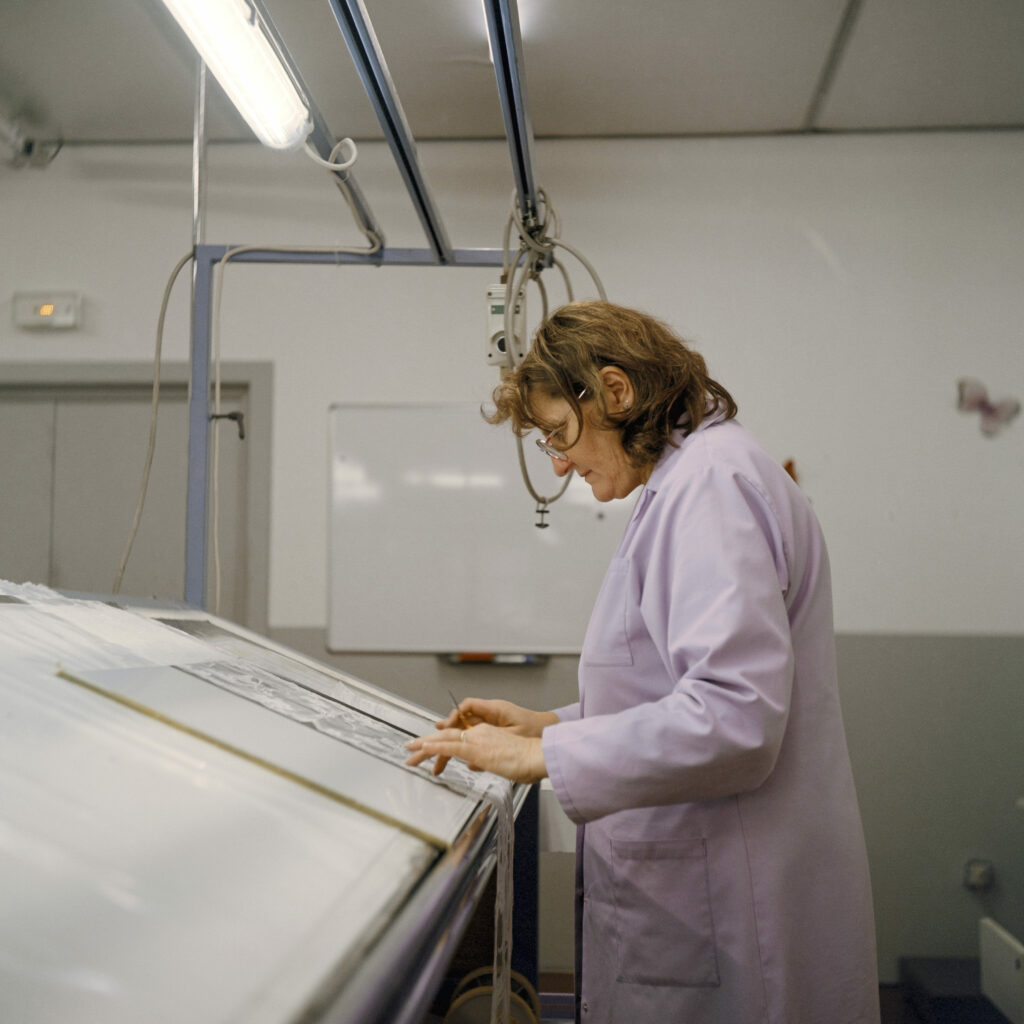 Photographie de Zélie Hallosserie / Light Motiv, issue de la série intitulée Dentelle Calais Caudry, représentant une femme en blouse blanche tournée vers la gauche et penchée sur une table blanche inclinée et regardant vers celle-ci, dans le cadre d'un reportage qui témoigne du travail et de la conception de la dentelle à Calais.