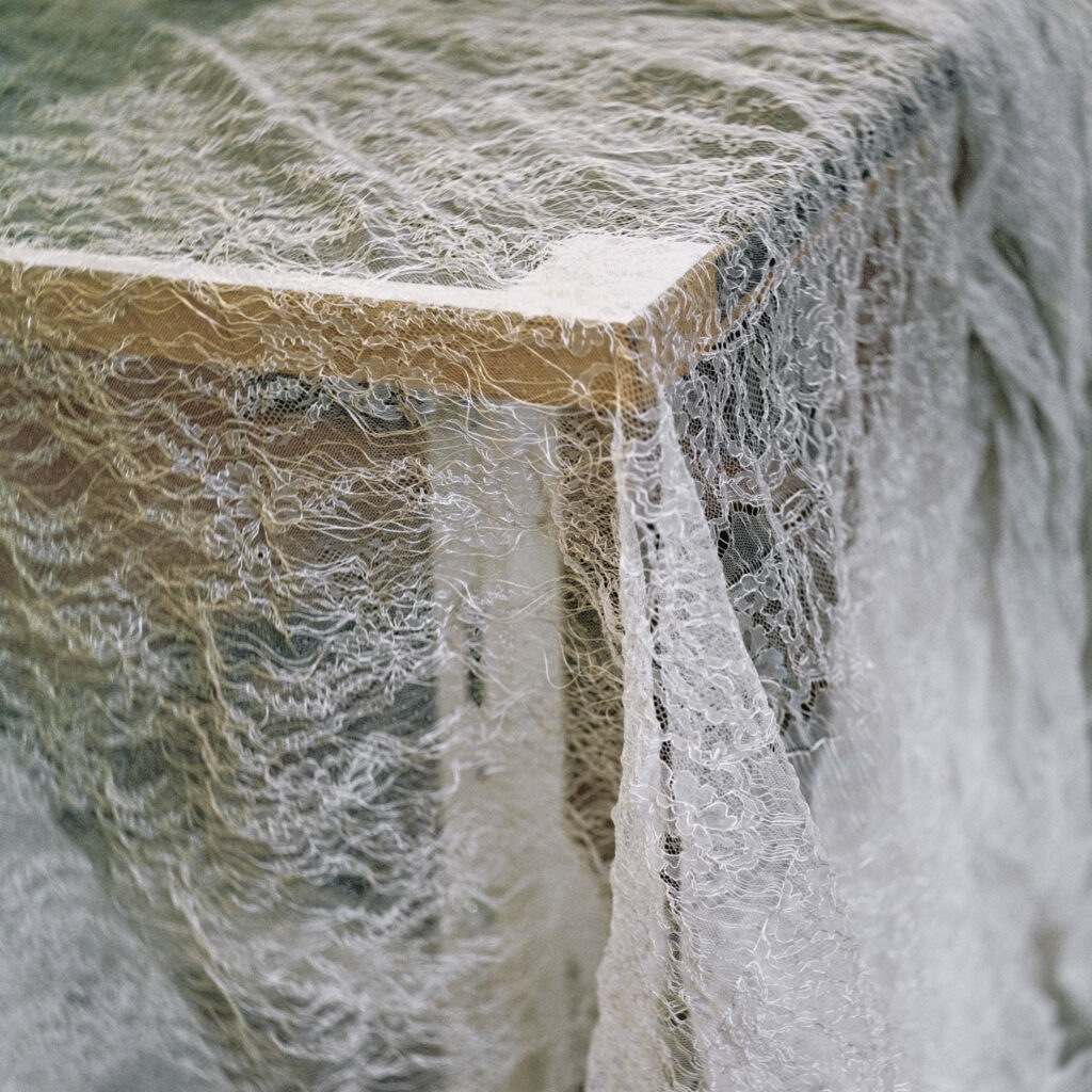 Photographie de Zélie Hallosserie / Light Motiv, issue de la série intitulée Dentelle Calais Caudry, représentant un grand tissu de dentelle blanche posé sur le coin d'une table en bois brun, dans le cadre d'un reportage qui témoigne du travail et de la conception de la dentelle à Calais.