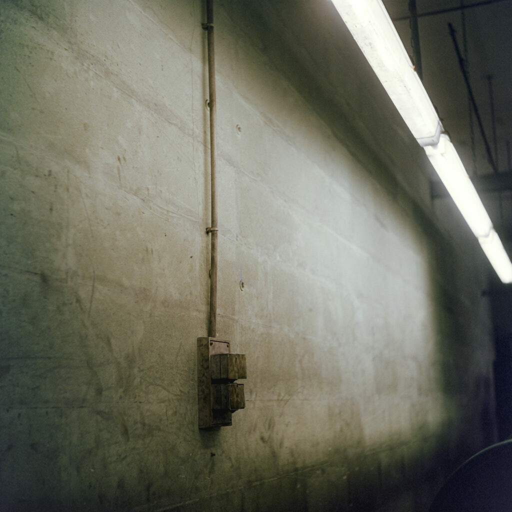 Photographie de Zélie Hallosserie / Light Motiv, issue de la série intitulée Dentelle Calais Caudry, représentant un mur en béton gris avec un boîtier le traversant surplombé d'une ligné de néons blancs, dans le cadre d'un reportage qui témoigne du travail et de la conception de la dentelle à Calais.