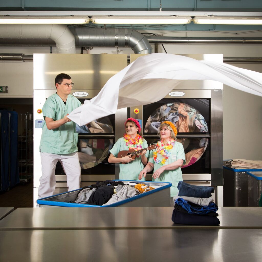 Photographie de Lionel Pralus / Light Motiv, issue de la série intitulée Désordre Ordinaire datant de 2020, représentant trois personnes dans une laverie, un homme sur la gauche tient un drap blanc en l'air dans ses mains pendant que deux femmes plus bas le regarde, dans le cadre d'un projet tourné autour de mise en scène photographiques avec les patients et professionnels du SAAS.