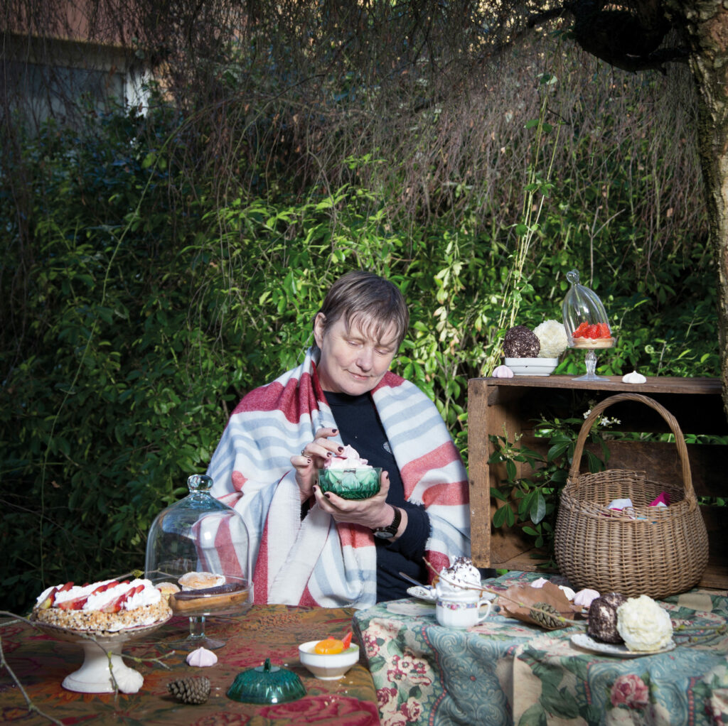 Photographie de Lionel Pralus / Light Motiv, issue de la série intitulée Désordre Ordinaire datant de 2020, représentant une femme assise en extérieur entourée de tables avec des pâtisseries dont une dans ses mains, dans le cadre d'un projet tourné autour de mise en scène photographiques avec les patients et professionnels du SAAS.