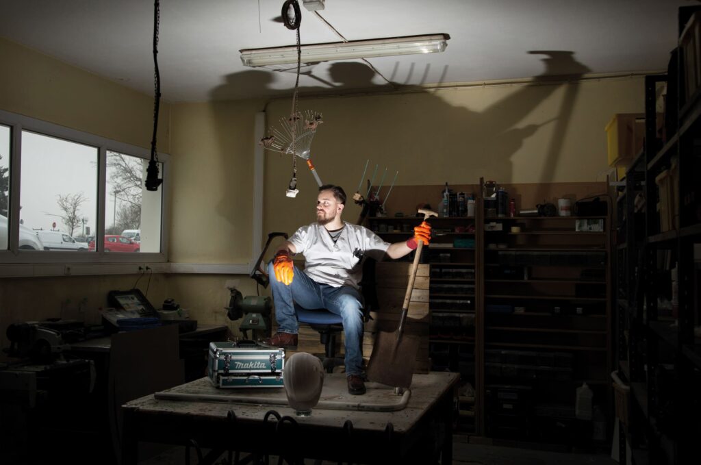 Photographie de Lionel Pralus / Light Motiv, issue de la série intitulée Désordre Ordinaire datant de 2020, représentant un homme assis sur une chaise sur une table tenant avec des gants de jardinages une grande pelle et éclairé par un projecteur par le bas, dans le cadre d'un projet tourné autour de mise en scène photographiques avec les patients et professionnels du SAAS.
