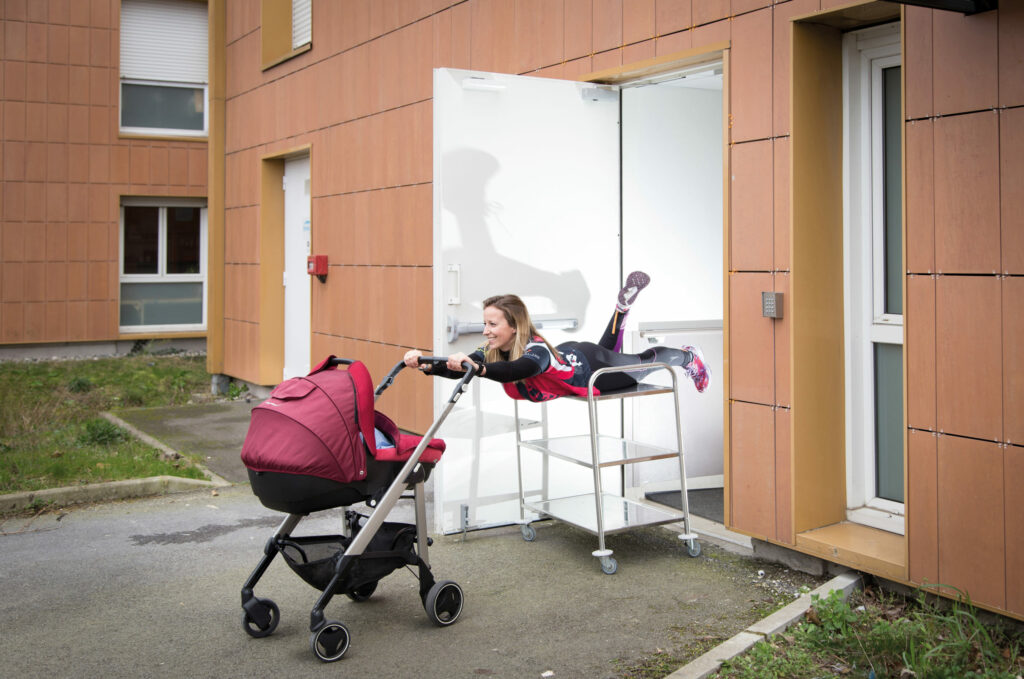 Photographie de Lionel Pralus / Light Motiv, issue de la série intitulée Désordre Ordinaire datant de 2020, représentant une femme souriante étendue sur une desserte dans l'entrebâillement d'une porte en tenant à bout de bras une poussette, dans le cadre d'un projet tourné autour de mise en scène photographiques avec les patients et professionnels du SAAS.