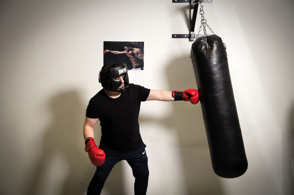 Photographie de Lionel Pralus / Light Motiv, issue de la série intitulée Désordre Ordinaire datant de 2020, représentant un homme debout en train de boxer un sac de boxe à l'aide de gants rouges et une image de Mohamed Ali accrochée au mur derrière lui, dans le cadre d'un projet tourné autour de mise en scène photographiques avec les patients et professionnels du SAAS.