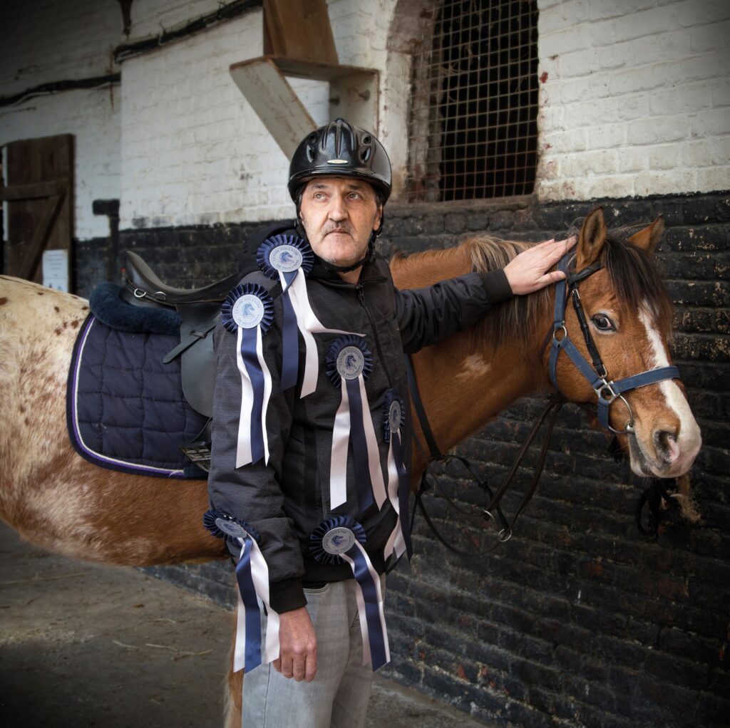 Photographie de Lionel Pralus / Light Motiv, issue de la série intitulée Désordre Ordinaire datant de 2020, représentant un homme debout carressant un cheval marron dans une écurie, dans le cadre d'un projet tourné autour de mise en scène photographiques avec les patients et professionnels du SAAS.
