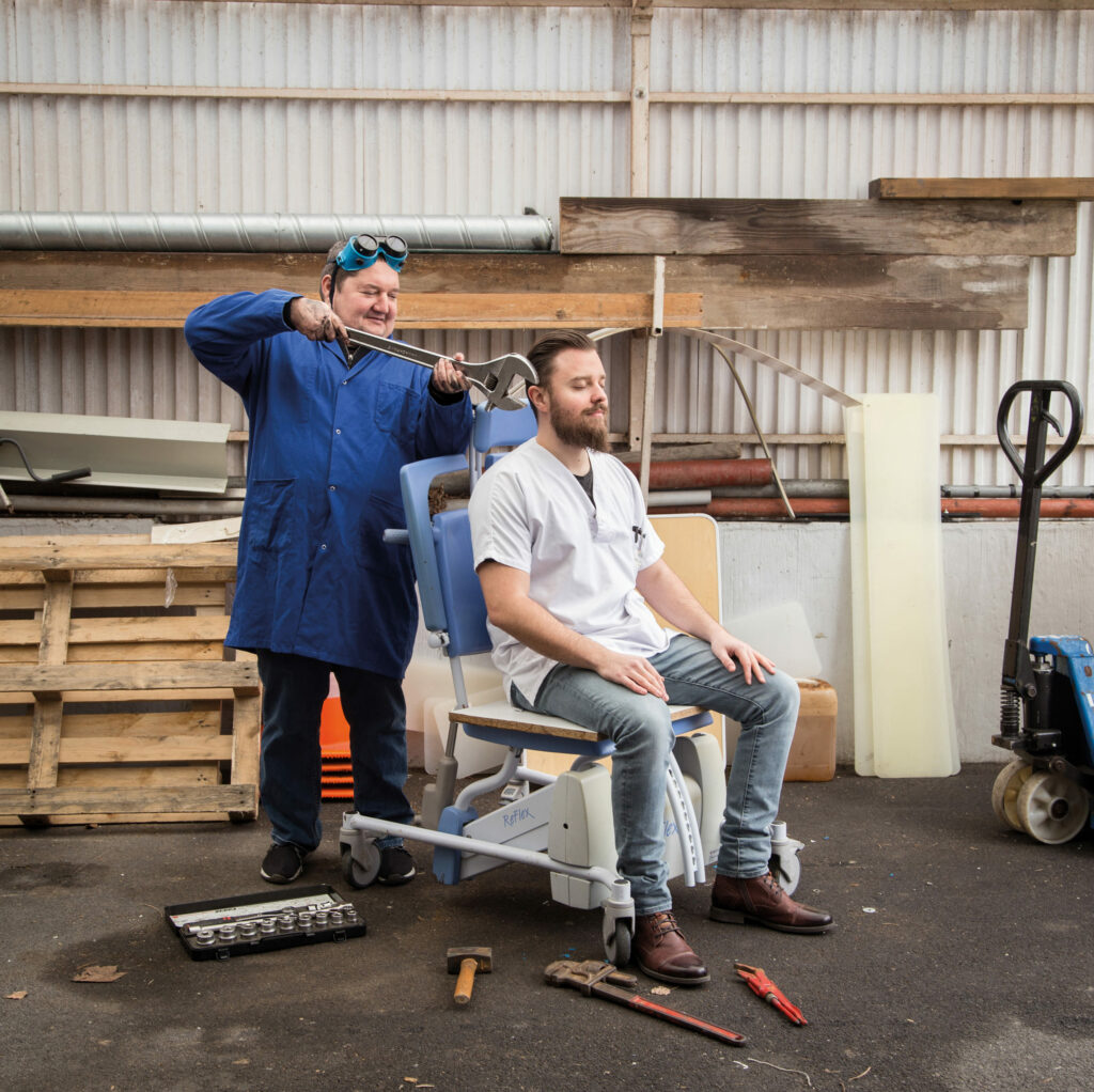 Photographie de Lionel Pralus / Light Motiv, issue de la série intitulée Désordre Ordinaire datant de 2020, représentant un homme en blouse blanche assis sur une chaise médicale et un autre homme en blouse bleue derrière lui occupé avec une grande clé à molette au niveau de son oreille, dans le cadre d'un projet tourné autour de mise en scène photographiques avec les patients et professionnels du SAAS.