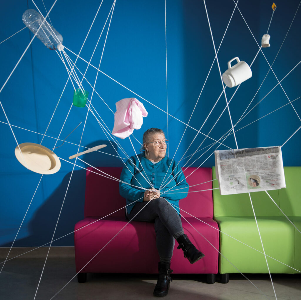 Photographie de Lionel Pralus / Light Motiv, issue de la série intitulée Désordre Ordinaire datant de 2020, représentant une femme assise sur un fauteuil rose dont des fils blancs s'échappent de part et d'autre de ses mains attachant divers objets en suspend, dans le cadre d'un projet tourné autour de mise en scène photographiques avec les patients et professionnels du SAAS.