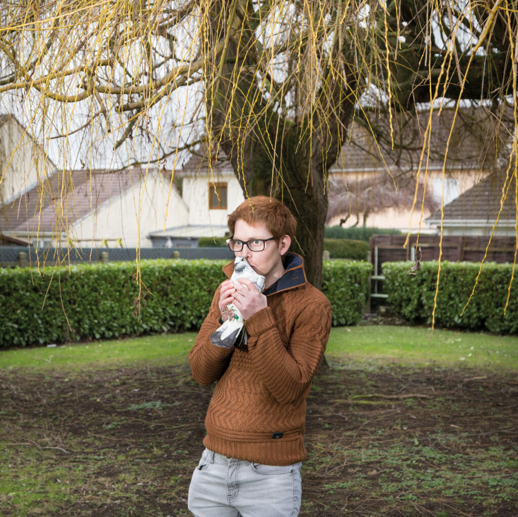 Photographie de Lionel Pralus / Light Motiv, issue de la série intitulée Désordre Ordinaire datant de 2020, représentant un homme debout devant un arbre tenant un pigeon blanc et gris dans ses deux mains jointes devant son visage, dans le cadre d'un projet tourné autour de mise en scène photographiques avec les patients et professionnels du SAAS.