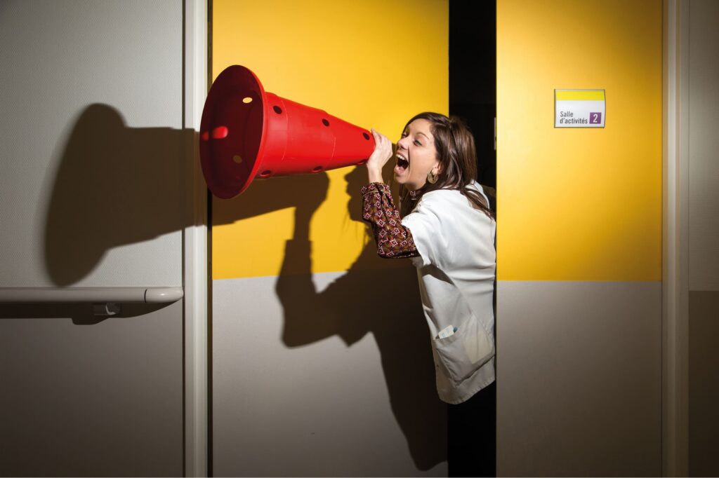 Photographie de Lionel Pralus / Light Motiv, issue de la série intitulée Désordre Ordinaire datant de 2020, représentant une jeune femme debout dans l'entrebâillement d'une porte avec un plot en guise de mégaphone dans sa main dirigé vers sa bouche, dans le cadre d'un projet tourné autour de mise en scène photographiques avec les patients et professionnels du SAAS.