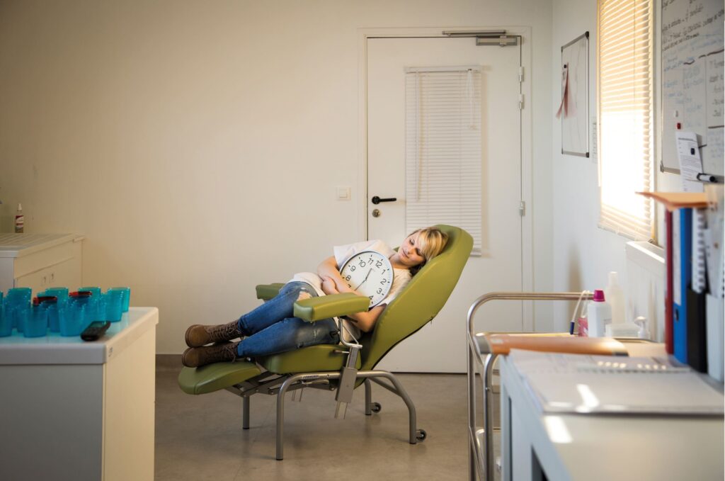 Photographie de Lionel Pralus / Light Motiv, issue de la série intitulée Désordre Ordinaire datant de 2020, représentant une femme en blouse blanche recroquevillée sur une chaise verte médicale tenant une horloge entre ses bras, dans le cadre d'un projet tourné autour de mise en scène photographiques avec les patients et professionnels du SAAS.
