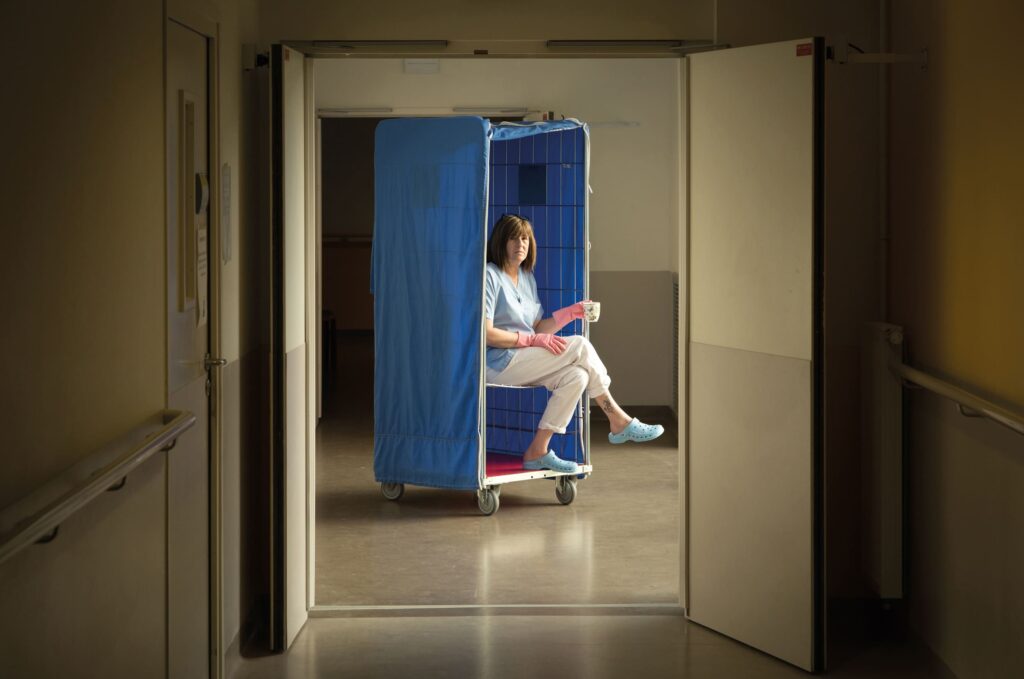 Photographie de Lionel Pralus / Light Motiv, issue de la série intitulée Désordre Ordinaire datant de 2020, représentant une femme assise sur un chariot métallique recouvert d'un tissu bleu dans un couloir en tenant une tasse dans sa main, dans le cadre d'un projet tourné autour de mise en scène photographiques avec les patients et professionnels du SAAS.