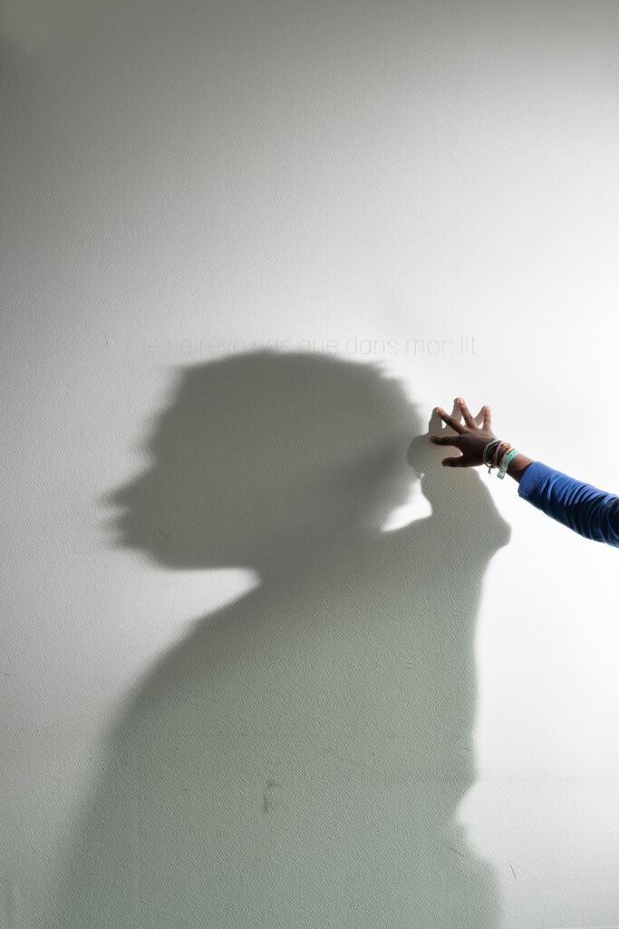 Photographie de Lionel Pralus / Light Motiv, issue de la série intitulée Le Jour La Nuit datant de 2024, représentant l'ombre d'une figure humaine projetée sur un mur blanc dont on voit la main posée sur le mur, dans le cadre d'un projet tourné autour de jeunes et de l'éclairage hospitalier.
