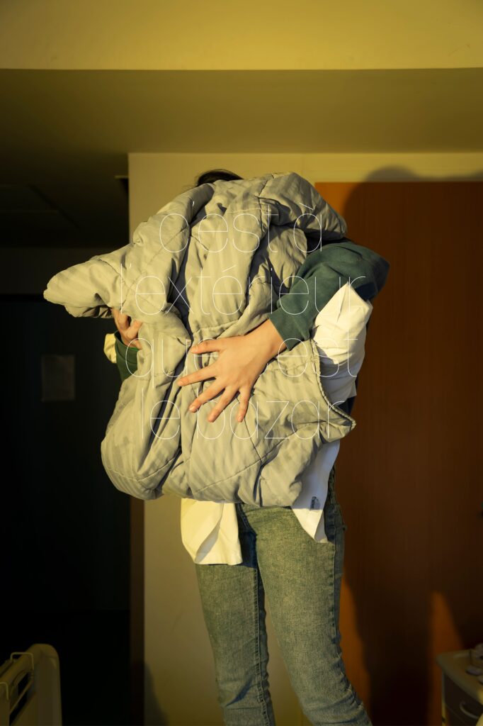 Photographie de Lionel Pralus / Light Motiv, issue de la série intitulée Le Jour La Nuit datant de 2024, représentant une personne tenant un ensemble de draps gris et blanc entre les mains dissimulant son visage avec un texte en filigrane "c'est à l'extérieur que c'est le bazar", dans le cadre d'un projet tourné autour de jeunes et de l'éclairage hospitalier.