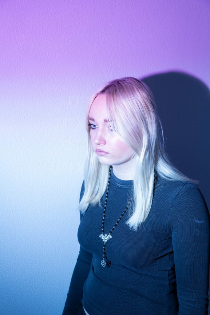 LP - LE JOUR LA NUIT Photographie de Lionel Pralus / Light Motiv, issue de la série intitulée Le Jour La Nuit datant de 2024, représentant une jeune fille tournée de trois quart debout devant un mur blanc avec une lumière bleue-violette dont le regard est tourné vers la gauche, dans le cadre d'un projet tourné autour de jeunes et de l'éclairage hospitalier.