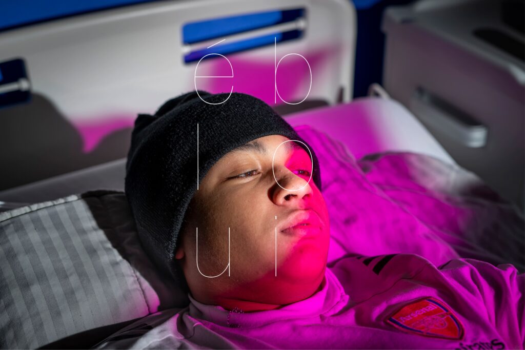 Photographie de Lionel Pralus / Light Motiv, issue de la série intitulée Le Jour La Nuit datant de 2024, représentant un plan rapproché sur le visage d'un jeune homme face caméra allongé sur un lit d'hôpital dont le regard est tourné vers la droite de l'image avec un texte en filigrane "ébloui", dans le cadre d'un projet tourné autour de jeunes et de l'éclairage hospitalier.