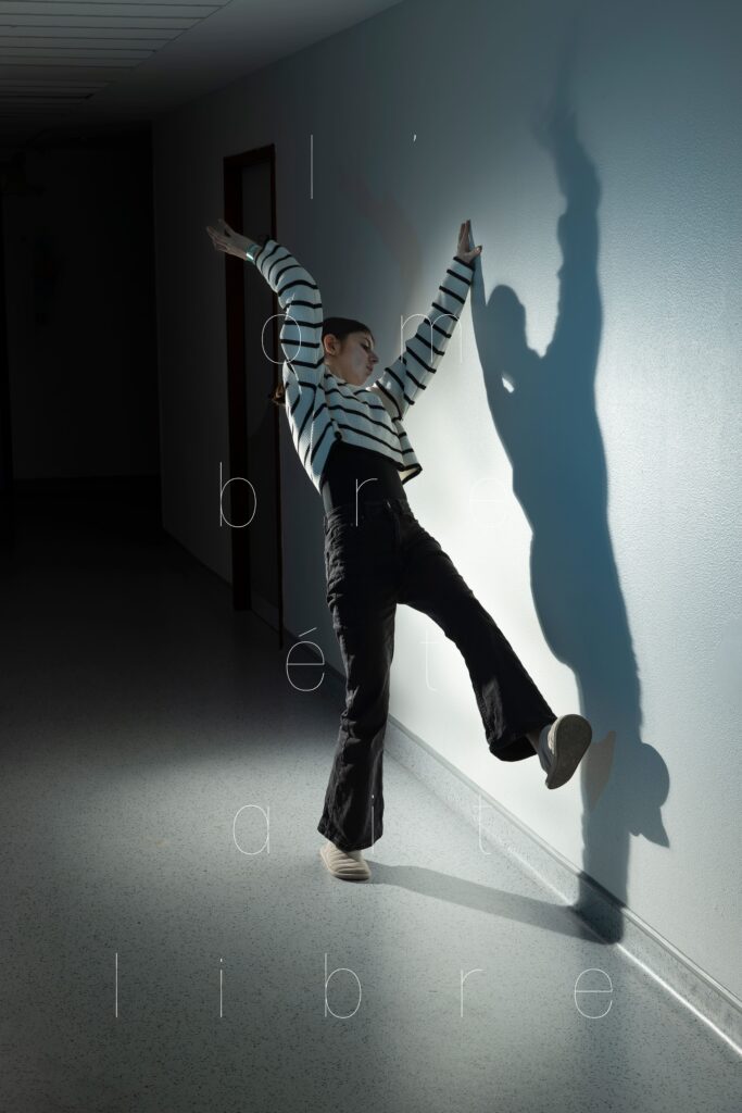 Photographie de Lionel Pralus / Light Motiv, issue de la série intitulée Le Jour La Nuit datant de 2024, représentant une jeune fille dansant dans un couloir d'hôpital avec une lumière blanche projetant son ombre sur le mur à côté d'elle avec un texte en filigrane "l'ombre était libre", dans le cadre d'un projet tourné autour de jeunes et de l'éclairage hospitalier.