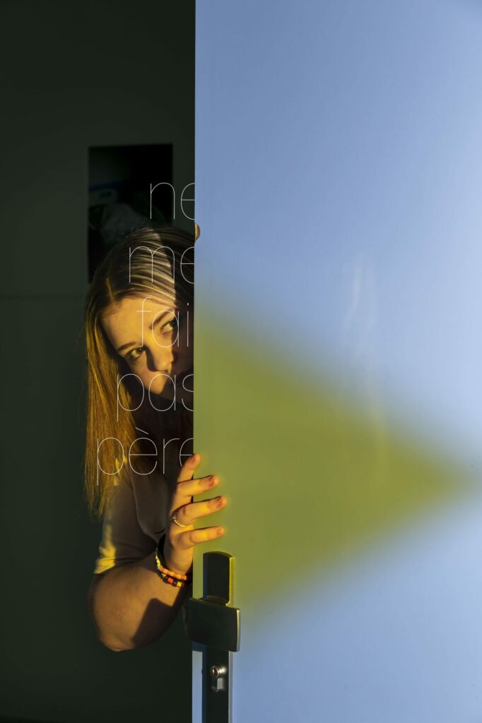 LP - LE JOUR LA NUIT Photographie de Lionel Pralus / Light Motiv, issue de la série intitulée Le Jour La Nuit datant de 2024, représentant une jeune fille cachée à moitié derrière une porte avec un texte en filigrane énigmatique, dans le cadre d'un projet tourné autour de jeunes et de l'éclairage hospitalier.