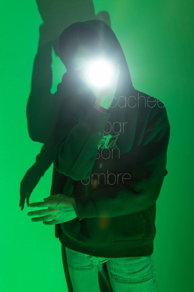LP - LE JOUR LA NUIT Photographie de Lionel Pralus / Light Motiv, issue de la série intitulée Le Jour La Nuit datant de 2024, représentant une personne debout contre un mur avec une source de lumière blanche tournée vers la caméra et un éclairage vert derrière elle avec un texte en filigrane "cachée par son ombre", dans le cadre d'un projet tourné autour de jeunes et de l'éclairage hospitalier.