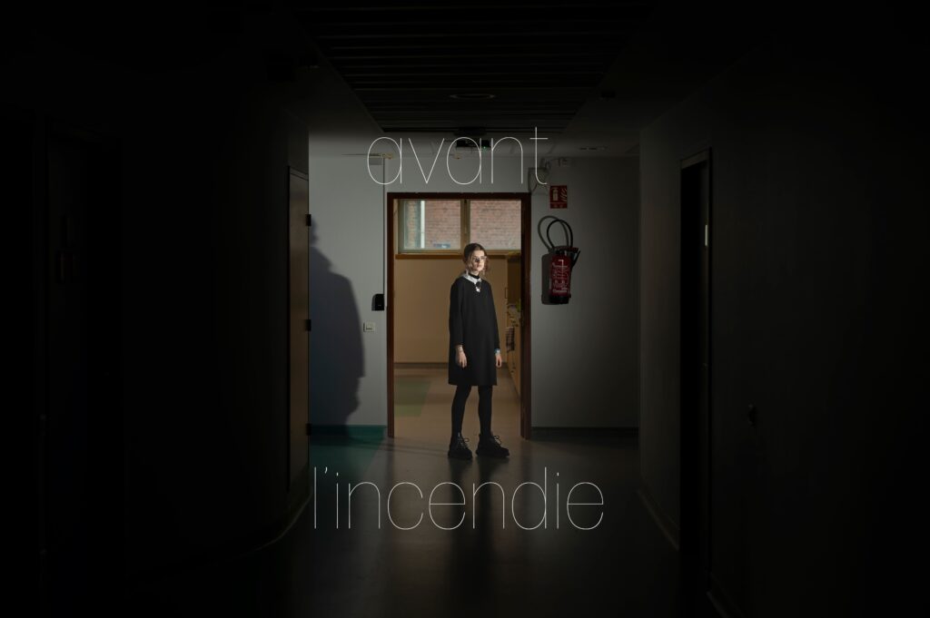 Photographie de Lionel Pralus / Light Motiv, issue de la série intitulée Le Jour La Nuit datant de 2024, représentant une jeune fille debout dans un couloir d'hôpital vide faiblement éclairé avec un texte en filigrane "avant l'incendie", dans le cadre d'un projet tourné autour de jeunes et de l'éclairage hospitalier.