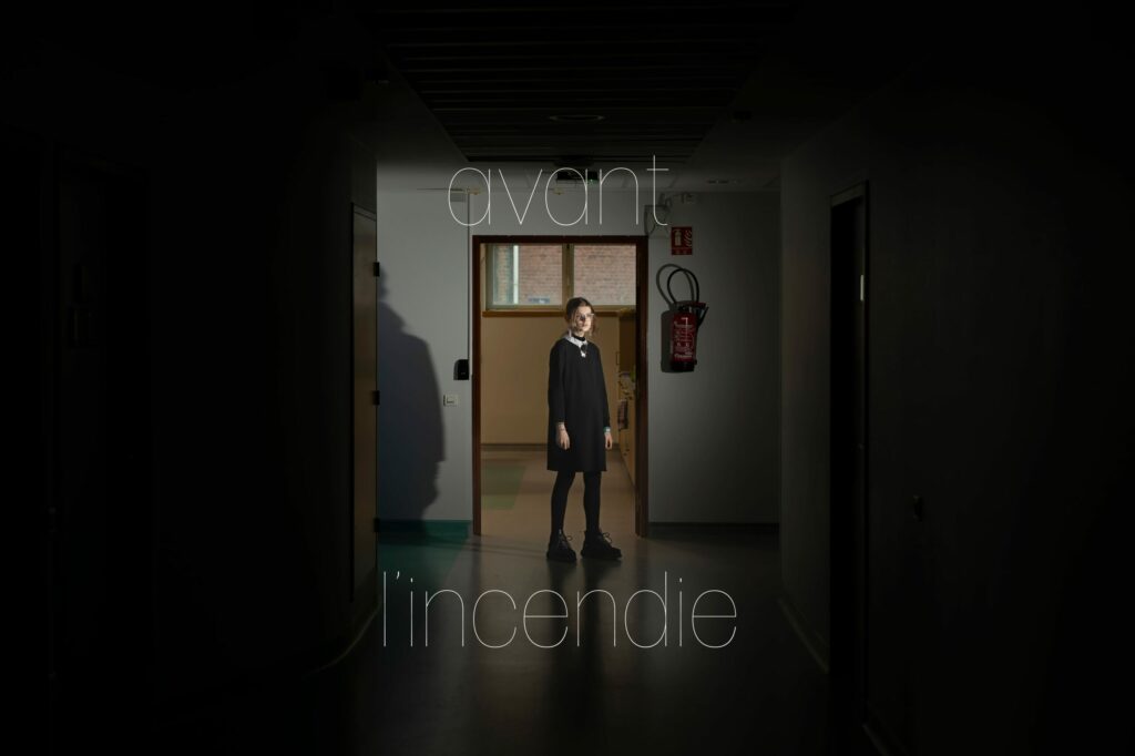 LP - LE JOUR LA NUIT Photographie de Lionel Pralus / Light Motiv, issue de la série intitulée Le Jour La Nuit datant de 2024, représentant une jeune fille debout dans un couloir d'hôpital vide faiblement éclairé avec un texte en filigrane "avant l'incendie", dans le cadre d'un projet tourné autour de jeunes et de l'éclairage hospitalier.