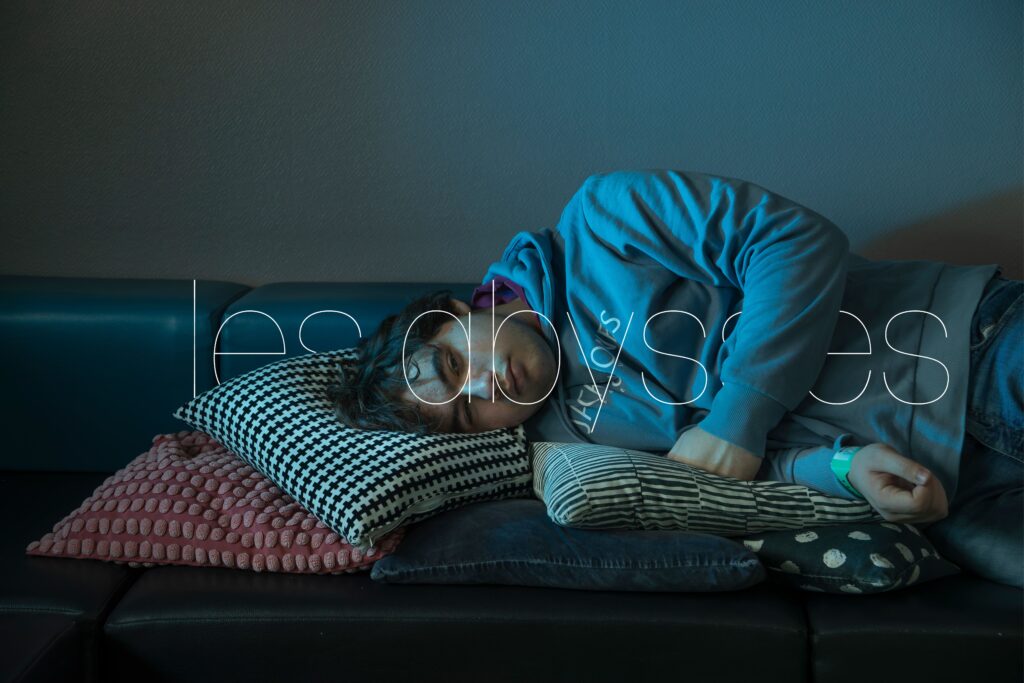 Photographie de Lionel Pralus / Light Motiv, issue de la série intitulée Le Jour La Nuit datant de 2024, représentant un jeune homme allongé sur un canapé et des coussins regardant vers la caméra avec un texte en filigrane "les abysses", dans le cadre d'un projet tourné autour de jeunes et de l'éclairage hospitalier.