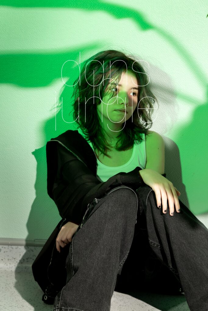 Photographie de Lionel Pralus / Light Motiv, issue de la série intitulée Le Jour La Nuit datant de 2024, représentant une jeune femme assise contre un mur éclairée d'une lumière verte et le visage tourné vers la droite avec un texte en filigrane "dans la jungle", dans le cadre d'un projet tourné autour de jeunes et de l'éclairage hospitalier.