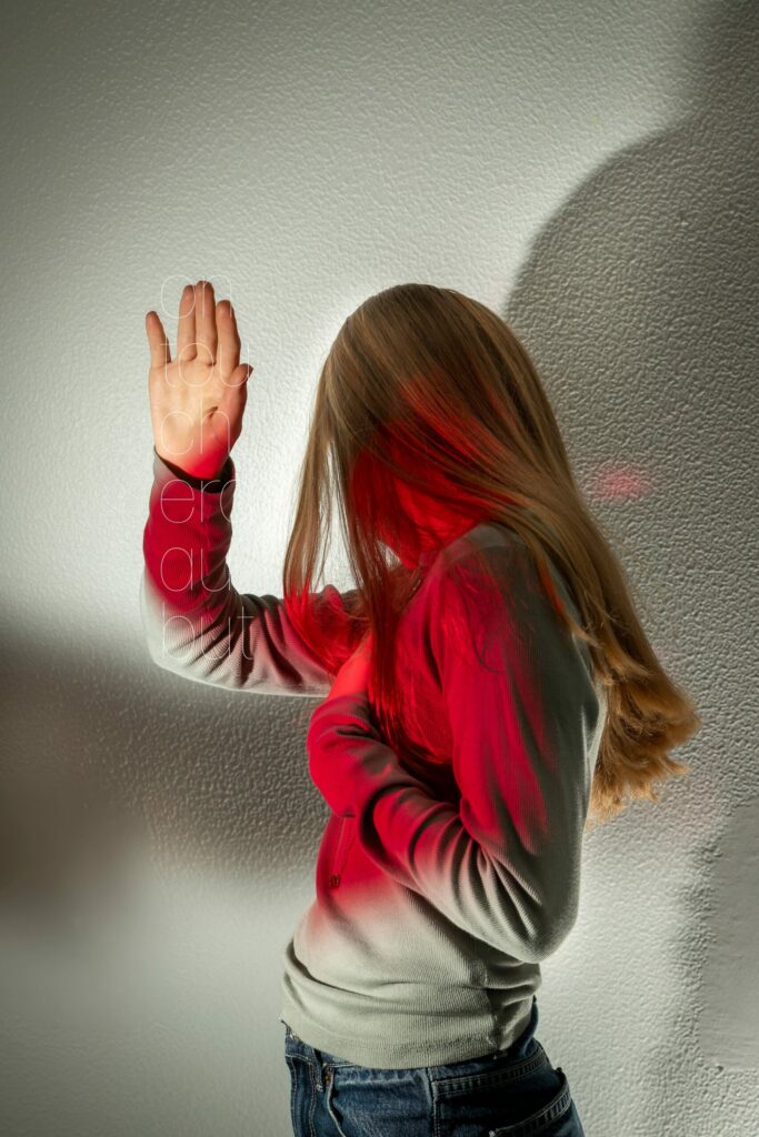 LP - LE JOUR LA NUIT Photographie de Lionel Pralus / Light Motiv, issue de la série intitulée Le Jour La Nuit datant de 2024, représentant une jeune fille le visage tourné derrière elle vers le mur avec la main droite levée en l'air et éclairée d'une lumière rouge vers sa poitrine avec un texte en filigrane "on touchera au but", dans le cadre d'un projet tourné autour de jeunes et de l'éclairage hospitalier.