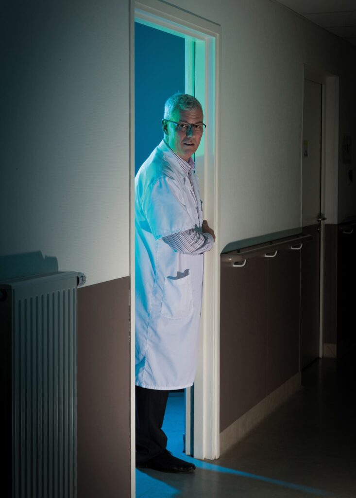 Photographie de Lionel Pralus / Light Motiv, issue de la série intitulée Désordre Ordinaire datant de 2020, représentant un homme dans l'encadrement d'une porte depuis une pièce dont une lumière bleue en ressort, dans le cadre d'un projet tourné autour de mise en scène photographiques avec les patients et professionnels du SAAS.